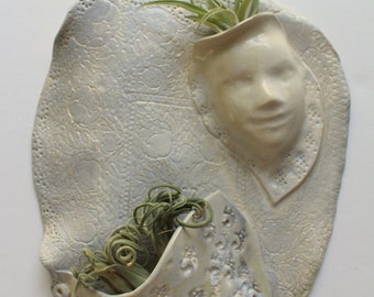 May include: White ceramic wall planter with two pockets, one shaped like a face and the other a simple pocket. Both pockets are filled with green air plants.