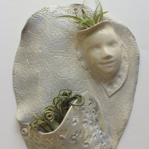 May include: White ceramic wall planter with two pockets, one shaped like a face and the other a simple pocket. Both pockets are filled with green air plants.