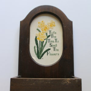May include: A wooden wall-mounted shelf with a framed cross-stitch design featuring yellow daffodils and the text "Take the time to smell the flowers".