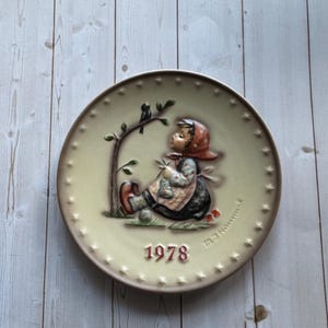 May include: A decorative plate with a relief image of a child knitting under a tree, with a bird perched on a branch. The plate has a cream-colored background, a brown border, and the year "1978" in red. The plate is round and has star-shaped accents.
