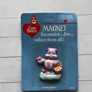 Könnte beinhalten: Ein Care Bears Share Bear Magnet auf einer blauen Karte. Der lila Bärenmagnet hat ein rosa Eis auf dem Bauch und hält einen Korb mit Herzen. Die Karte lautet "MAGNET Decorative...fun... collect them all!"