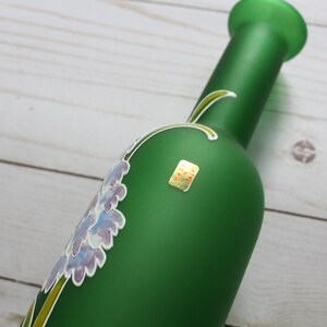 Puede incluir: Una botella de vidrio verde con un dise&ntilde;o floral en tonos morados y blancos. La botella tiene un cuello largo y una peque&ntilde;a etiqueta. El dise&ntilde;o floral est&aacute; pintado en la botella.