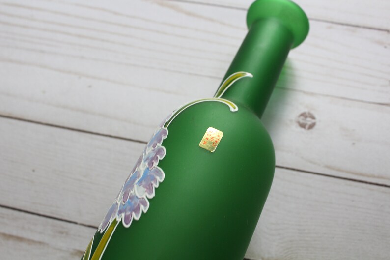 Puede incluir: Una botella de vidrio verde con un dise&ntilde;o floral en tonos morados y blancos. La botella tiene un cuello largo y una peque&ntilde;a etiqueta. El dise&ntilde;o floral est&aacute; pintado en la botella.