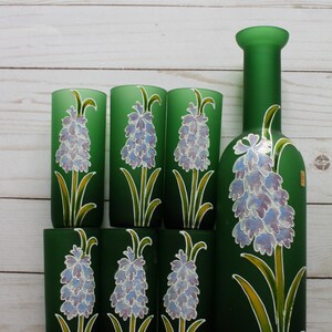 Puede incluir: Una botella de vidrio verde y seis vasos a juego. Cada pieza est&aacute; decorada con un dise&ntilde;o floral pintado a mano con una flor de lavanda con hojas y tallos verdes. El conjunto se exhibe sobre una superficie de madera clara.