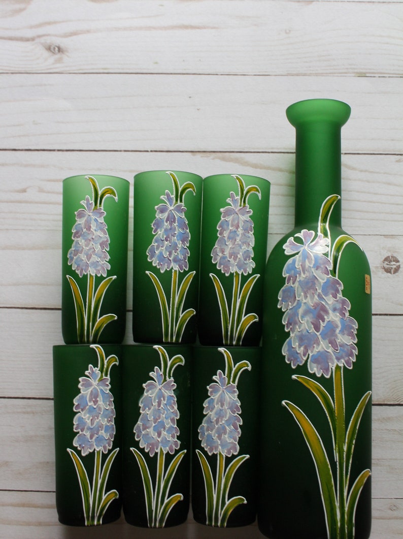 Puede incluir: Una botella de vidrio verde y seis vasos a juego. Cada pieza est&aacute; decorada con un dise&ntilde;o floral pintado a mano con una flor de lavanda con hojas y tallos verdes. El conjunto se exhibe sobre una superficie de madera clara.