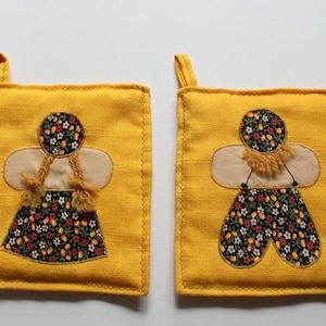 Op de afbeelding: Twee gele ovenwanten met een bloemenpatroon. Elke ovenwant heeft een gestikte pop met een bloemenjurk en een bruin haarbandje.