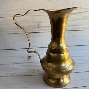 May include: A vintage brass pitcher with a curved handle and a flared spout. The pitcher has a bulbous body with horizontal ridges and a pedestal base. The brass has a warm, golden hue, and the pitcher is set against a light-colored wooden background.