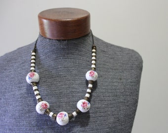 May include: A white porcelain necklace with pink and blue floral accents. The necklace features round beads with a floral design and is strung on a brown cord.