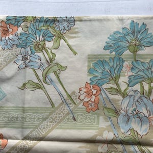 Op de afbeelding: Crèmekleurige stof met een bloemenpatroon. Grote blauwe en perzikbloemen, groene bladeren en decoratieve geometrische randen zijn zichtbaar. Het ontwerp doet denken aan vintage textiel, geschikt voor woondecoratie of knutselprojecten.