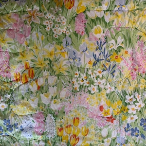 以下が含まれることがあります： 黄色、ピンク、白、青の色合いの花が緑色の背景に描かれた、花のキルト。キルトはキルティングパターンで、デザインに質感を加えています。花には、チューリップ、水仙、ユリが含まれています。