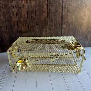 May include: A gold-colored metal tissue box cover. The rectangular box has a mesh design with floral embellishments, including roses and leaves. The top features a rectangular opening for tissue dispensing.