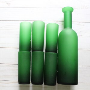 Puede incluir: Un conjunto de siete art&iacute;culos de vidrio verde, que incluyen seis vasos cil&iacute;ndricos y una botella. Los vasos y la botella tienen un acabado esmerilado, y el color pasa de un verde m&aacute;s claro en la parte superior a un verde m&aacute;s oscuro en la parte inferior.