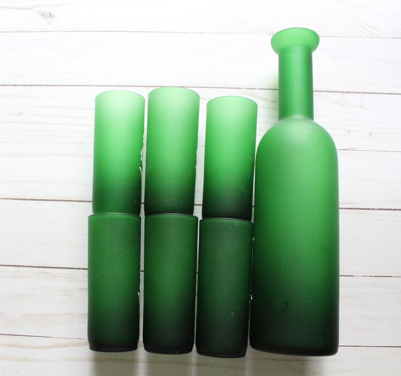 Puede incluir: Un conjunto de siete art&iacute;culos de vidrio verde, que incluyen seis vasos cil&iacute;ndricos y una botella. Los vasos y la botella tienen un acabado esmerilado, y el color pasa de un verde m&aacute;s claro en la parte superior a un verde m&aacute;s oscuro en la parte inferior.
