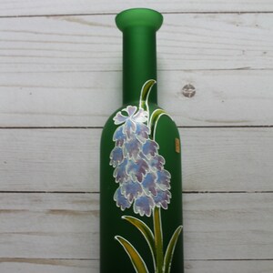 Puede incluir: Una botella de vidrio verde con un dise&ntilde;o floral pintado. La botella tiene un cuello largo y un cuerpo redondeado. El dise&ntilde;o floral es un racimo de flores azules y moradas con hojas y tallos verdes.