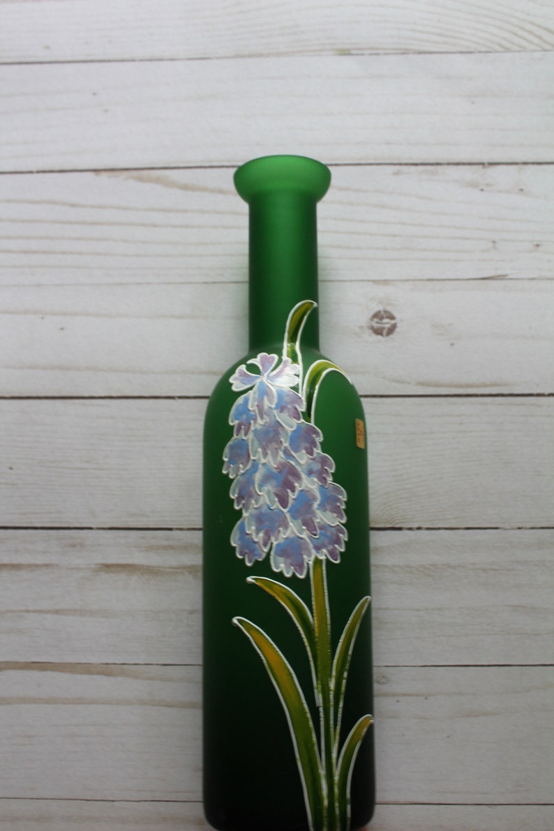 Puede incluir: Una botella de vidrio verde con un dise&ntilde;o floral pintado. La botella tiene un cuello largo y un cuerpo redondeado. El dise&ntilde;o floral es un racimo de flores azules y moradas con hojas y tallos verdes.