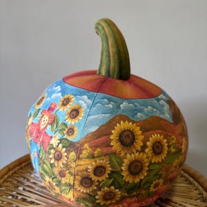 May include: A hand-painted fabric pumpkin with a fall scene featuring a scarecrow, sunflowers, and a mountain range. The pumpkin is orange with a green stem and is decorated with a blue sky and white clouds.