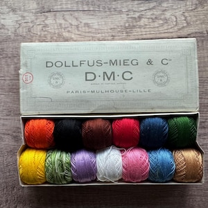 May include: A vintage DMC embroidery floss set in a cardboard box. The box lid is open, revealing a variety of colorful thread spools. The box is labeled "DOLLFUS-MIEG & C° D.M.C PARIS-MULHOUSE-LILLE". The spools are in shades of red, black, brown, pink, blue, green, yellow, and purple.