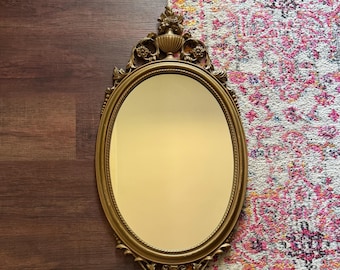 May include: An oval wall mirror with a gold-toned frame featuring ornate decorative carvings at the top and bottom. The frame includes a rope-like inner border and a central urn motif surrounded by floral elements at the apex.