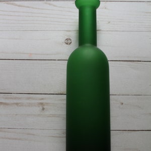Puede incluir: Una botella de vidrio verde con un acabado mate. La botella tiene un cuerpo largo y cil&iacute;ndrico y un cuello corto y redondeado. La botella est&aacute; vac&iacute;a y se encuentra sobre una superficie de madera blanca.
