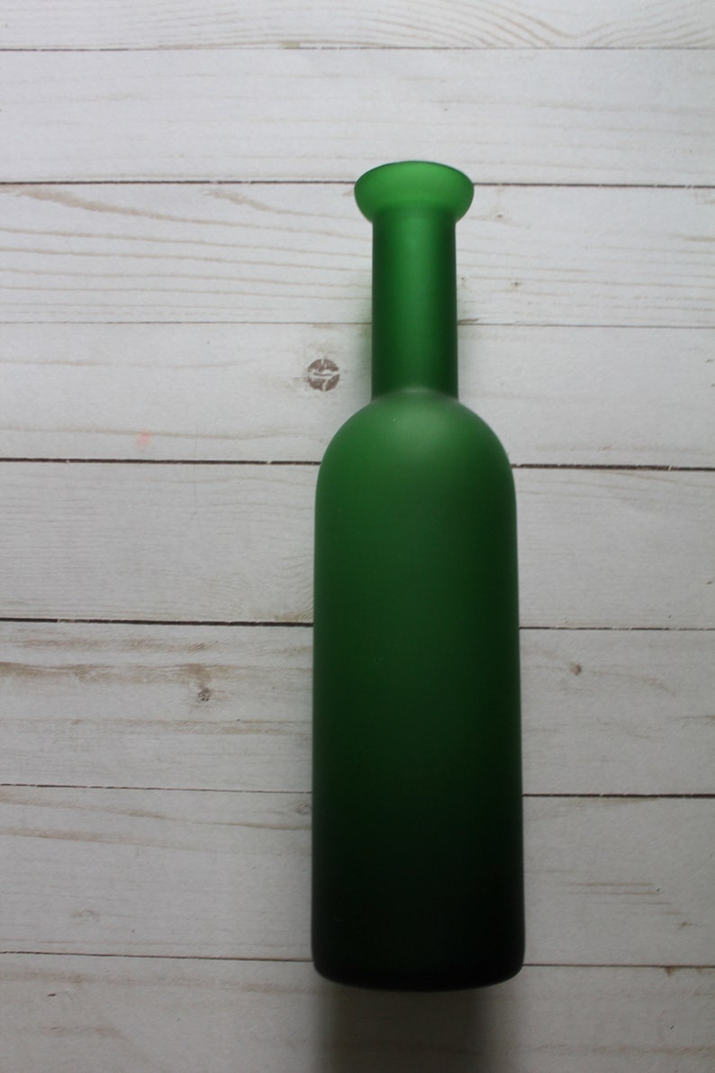 Puede incluir: Una botella de vidrio verde con un acabado mate. La botella tiene un cuerpo largo y cil&iacute;ndrico y un cuello corto y redondeado. La botella est&aacute; vac&iacute;a y se encuentra sobre una superficie de madera blanca.