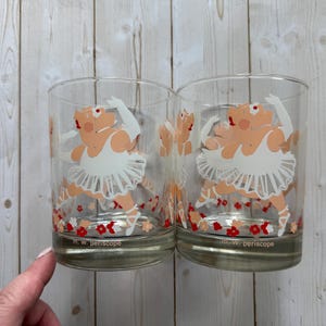 Puede incluir: Dos vasos transparentes con un diseño caprichoso de un cerdo con tutú blanco, zapatillas de ballet y guantes. El cerdo está en una pose de ballet. La base de los vasos tiene un estampado floral con flores rojas. El texto "m. w. periscope" está impreso en la base.