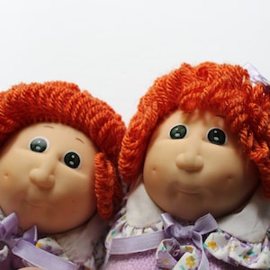 Może przedstawiać: Dwie lalki Cabbage Patch Kids z czerwonymi włosami z włóczki w fioletowych i białych sukienkach z kwiatowymi wzorami.
