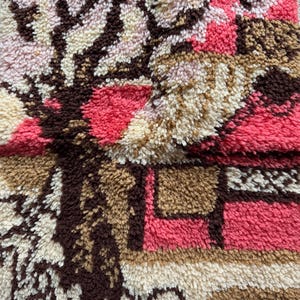 May include: Close-up of a textured rug with a floral design. The rug features a combination of colors, including pink, brown, beige, and white. The design appears to be a stylized depiction of flowers and branches.