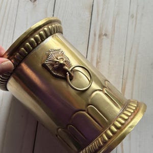 Puede incluir: Un jarrón cilíndrico de metal dorado con un diseño de cabeza de león y un asa de anillo. El jarrón tiene un patrón decorativo alrededor de la parte superior e inferior.