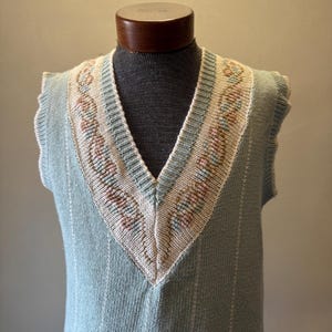 May include: A light blue knitted vest with a V-neck design. The neckline features a cream-colored border with pink and brown floral embroidery. Vertical white stripes run down the front of the vest, adding a subtle pattern. The vest is displayed on a mannequin.