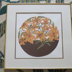 May include: A framed print of a circular arrangement of orange lilies with green leaves on a brown background. The print is signed "Margaret  W.  Bailey".