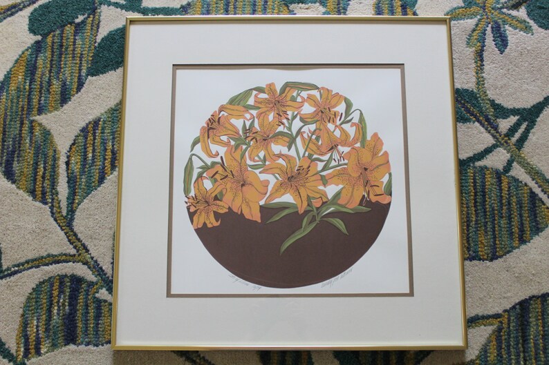 May include: A framed print of a circular arrangement of orange lilies with green leaves on a brown background. The print is signed "Margaret  W.  Bailey".