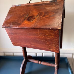 Puede incluir: Una caja de costura de madera vintage con tapa con bisagras y asa de latón. La caja está teñida de color marrón rojizo y está montada sobre un soporte de madera. La caja tiene una estética clásica y antigua.