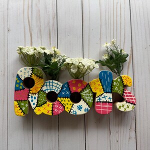May include: A wooden sign spelling "roots" with each letter painted in a patchwork style with different colors and patterns. The sign is decorated with white flowers.