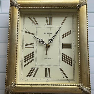 May include: A rectangular Bulova quartz wall clock with a gold-toned frame featuring decorative corner accents. The cream-colored face displays black Roman numerals, thin black hands, and a minute track around the perimeter of the clock dial.