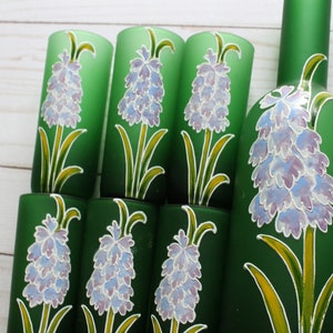 Puede incluir: Un conjunto de vasos de vidrio verde y una botella, todos decorados con un dise&ntilde;o floral. Las flores son de color p&uacute;rpura claro y blanco, con tallos y hojas amarillas y verdes. La botella y los vasos est&aacute;n sobre una superficie de madera clara.
