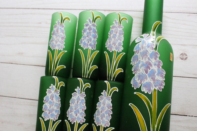 Puede incluir: Un conjunto de vasos de vidrio verde y una botella, todos decorados con un dise&ntilde;o floral. Las flores son de color p&uacute;rpura claro y blanco, con tallos y hojas amarillas y verdes. La botella y los vasos est&aacute;n sobre una superficie de madera clara.