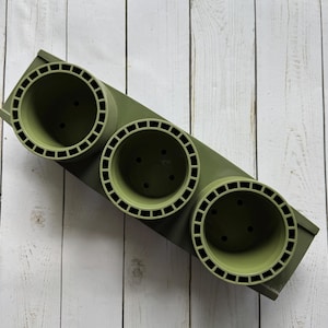 Puede incluir: Una jardinera rectangular verde oliva con tres aberturas circulares. Cada abertura tiene un borde decorativo con un patrón cuadrado. La jardinera está diseñada para contener varias plantas y está hecha de un material duradero.