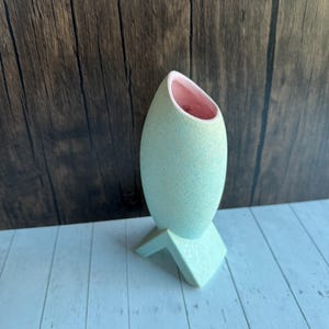 Puede incluir: Un jarrón de cerámica azul claro con una textura moteada y un interior rosa. El jarrón tiene forma ovalada y se asienta sobre una base geométrica. El fondo es una pared de madera y una superficie de madera blanca.