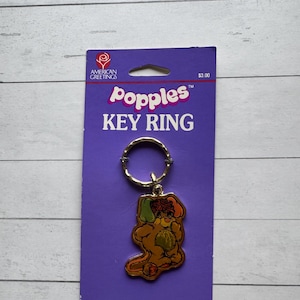 May include: A vintage Popples key ring on a purple card. The key ring features a gold-colored metal ring and a charm depicting a cartoon Popple character in shades of brown, orange, and green. The card reads "Popples KEY RING".