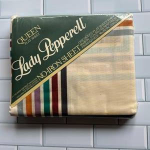 May include: A packaged Lady Pepperell queen flat sheet with a no-iron finish. The packaging features a dark green label with white text and a striped pattern in shades of cream, brown, teal, and burgundy. The finished size is 90x102 inches.