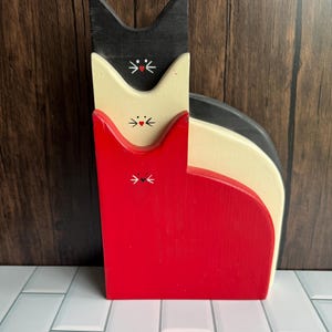 May include: A set of three nested wooden cat figures in black, cream, and red. Each cat features a painted face with a small red heart nose and white whiskers. The figures are stacked vertically, with the black cat on top, cream in the middle, and red at the base, creating a layered silhouette.
