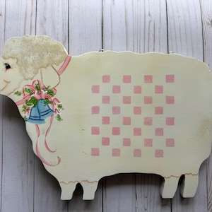 May include: A wooden sheep-shaped wall hanging with a cream-colored body and pink checkered pattern. The sheep has a fluffy head, a pink ribbon, and a floral and bell decoration. The background is a light wood.