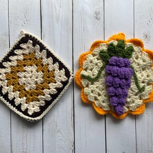 Puede incluir: Dos agarraderas de olla de ganchillo, una con un patrón cuadrado en marrón, blanco y amarillo, y la otra con un patrón redondo en amarillo, blanco y morado, con un diseño de uva.
