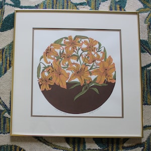 May include: A framed print of a stylised illustration of orange lilies in a brown vase. The print is titled "Lily in Bloom" and signed by the artist.