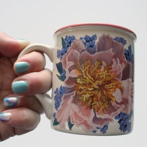 Peut inclure: Une tasse en céramique blanche avec un bord rose et un motif floral. Le motif représente un grand pivoine rose avec un centre jaune et des fleurs bleues qui l'entourent.