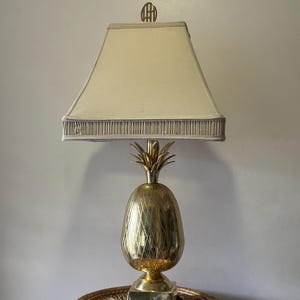 Puede incluir: Una lámpara de mesa de piña dorada con una pantalla blanca. La pantalla tiene un borde dorado y un remate dorado.
