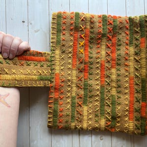 Pode incluir: Têxtil tecido em tons de ouro, laranja e verde, com um padrão repetitivo de listras verticais e bordados em ponto cruz. O têxtil é segurado por uma mão, mostrando sua textura e design.