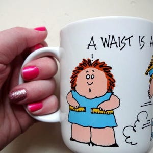 Op de afbeelding: Witte keramische mok met een blauwe, roze en zwarte cartoonillustratie. De mok heeft een zwart handvat en de tekst "A Waist Is A ..." staat op de voorkant.