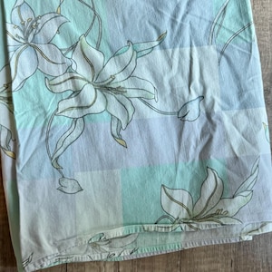 May include: A fabric textile featuring a light blue, mint green, and white checkered background pattern overlaid with large, outlined floral lily illustrations in dark green lines. The material has a soft, folded appearance with visible seams.