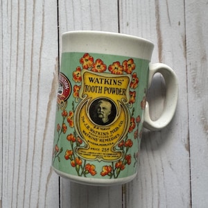 May include: Vintage Watkins' Tooth Powder mug. The mug features a light green background with orange floral accents and the text "Watkins' Tooth Powder." A portrait of a man is in the center. The mug has a white handle and rim.